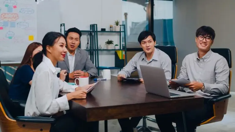 Five people sit around a conference table with papers and a laptop, appearing to discuss or present in an office meeting room.