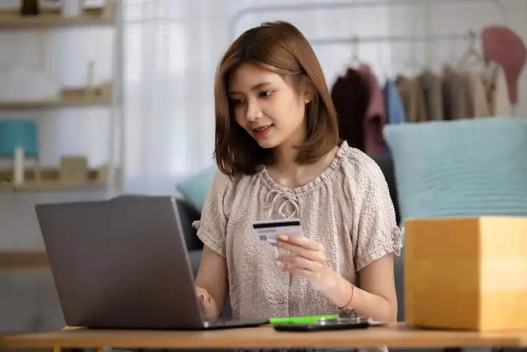 A woman sits at a table using a laptop and holding a credit card, with a cardboard box, green smartphone, and paperwork nearby.