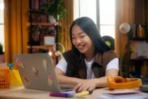 A person sits at a desk and smiles while using a laptop decorated with colorful stickers. The workspace includes pens, headphones, and shelves with plants in the background.
