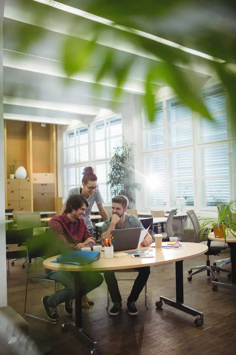 Three people collaborate at a round table in a bright, modern office space, with plants partially visible in the foreground.