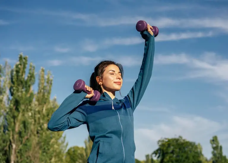 Woman in a blue jacket lifts purple dumbbells outdoors, with trees and blue sky in the background.